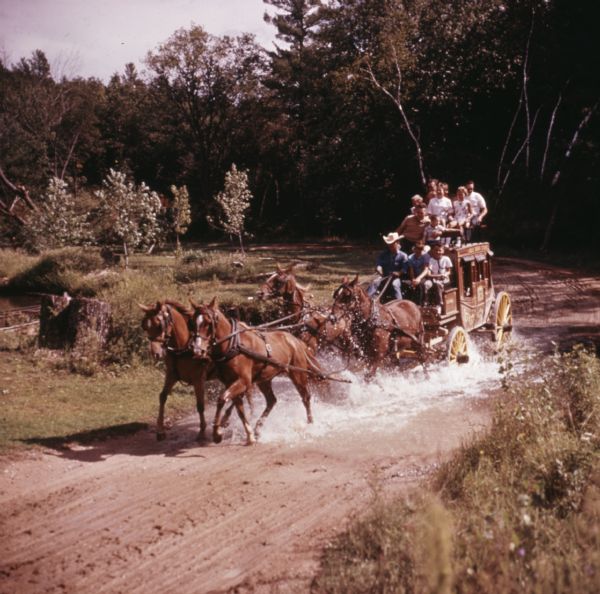 Slightly elevated view of four horses pulling a stagecoach through a puddle on a dirt road. A group of children and adults are sitting on top of the stagecoach as a man in a cowboy hat guides the horses.