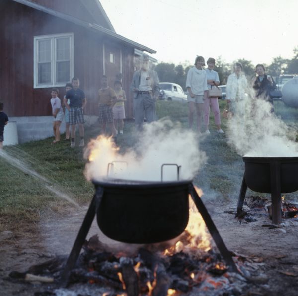 A crowd of men, women, and children are watching as two pit fires heat two large pots in the foreground. Someone (out of frame) is spraying water from the left to douse the fire.