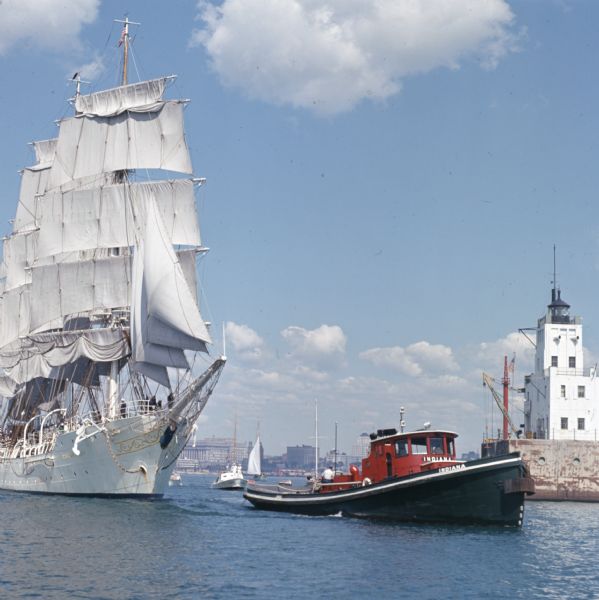 View across water towards the tugboat "Indiana" pulling the full-rigged, three-masted Norwegian ship "Christian Radich" into port. The sails and hull of the ship are white, with some gold decoration on the hull.
