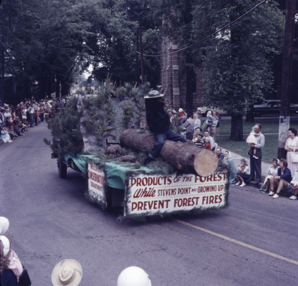 Elevated view over a crowd of people watching a parade. The Wisconsin Conservation Department float is coming down the road. It features a forest with taxidermied animals, and a person in a Smokey the Bear costume sitting on a log. A sign on the front of the float reads: "Products of the Forest, While Stevens Point was Growing Up, 'Prevent Forest Fires.'" A sign on the side of the float reads: "Wisconsin Conservation Department, Forest Protection Division."