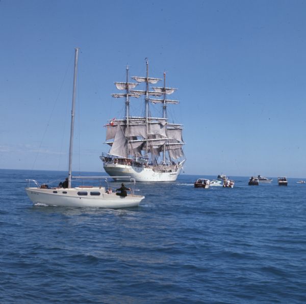 Nine boats are on the waters of Lake Michigan as they observe the "Christian Radich" sailing. The ship is a full-rigged, three-masted Norwegian ship. The sails and hull of the ship are white, with some gold decoration on the hull. A Norwegian flag is flying from the driver at the stern of the ship.