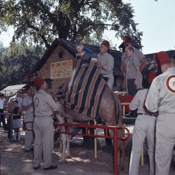 Men wearing red fez hats and khaki shirts and pants (the Zor Camel Patrol) are helping a boy get on a camel for a ride around Vilas Park.