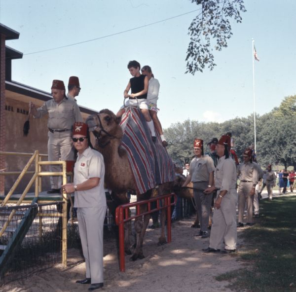 Men of the Zor Camel Patrol in Vilas Park are holding the reins of a camel as a boy and girl are sitting on the camel for a ride. A line of people are standing in the background. 