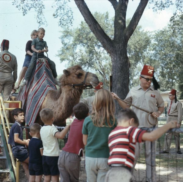 Children are waiting in a line behind a fence for their turn to ride a camel. Two children are sitting on a camel as men of the Zor Camel Patrol are leading the camel through Vilas Park.