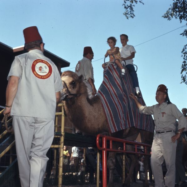 Low angle view of two children riding a double humped camel at the Henry Vilas Zoo. Men from the Zor Camel Patrol are wearing red fez hats and khaki shirts and pants and are preparing to guide the camel. 