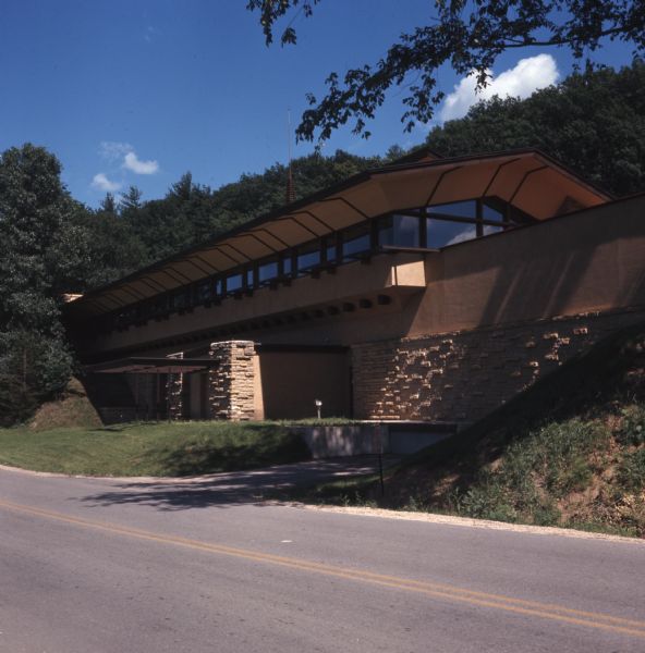 View of a brick and stucco building on the side of a road. It has an overhanging roof line and a cantilevered canopy over the lower entrance.