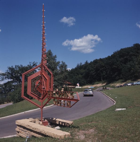A car is driving up an inclined road to a parking lot. A red metal sculptural sign in the foreground reads: "The Spring Green."