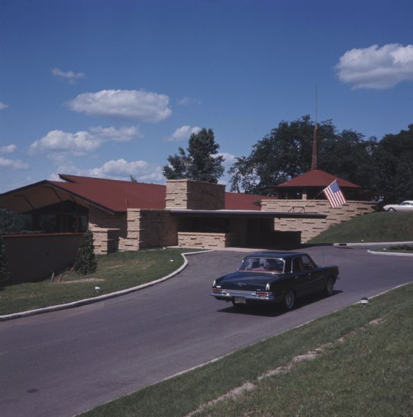 A car is driving up the road to The Spring Green, which has a brick and stucco building with a red roof. An American flag is flying from the canopy above the upper entrance.