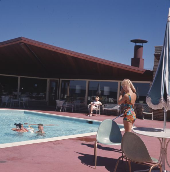 A woman in a bathing suit is standing on the edge of a swimming pool and taking a photograph of two children swimming.