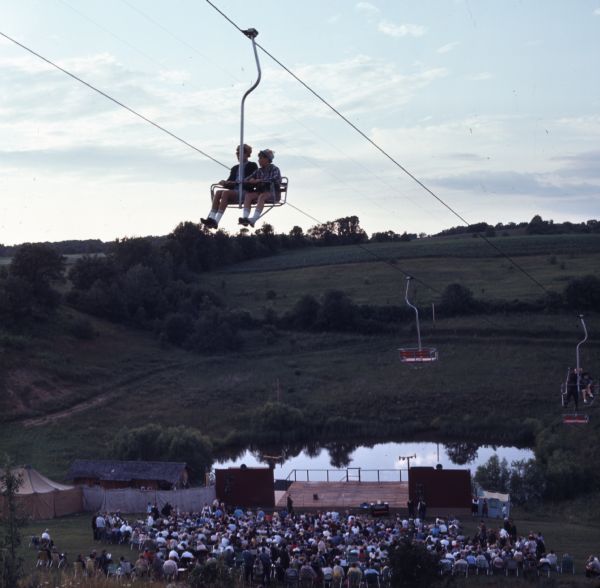 Elevated view of two boys riding a ski lift high above a crowd of people and a stage below them a the bottom of a hill. The crowd is waiting for the musical "Song of Norway" to start.