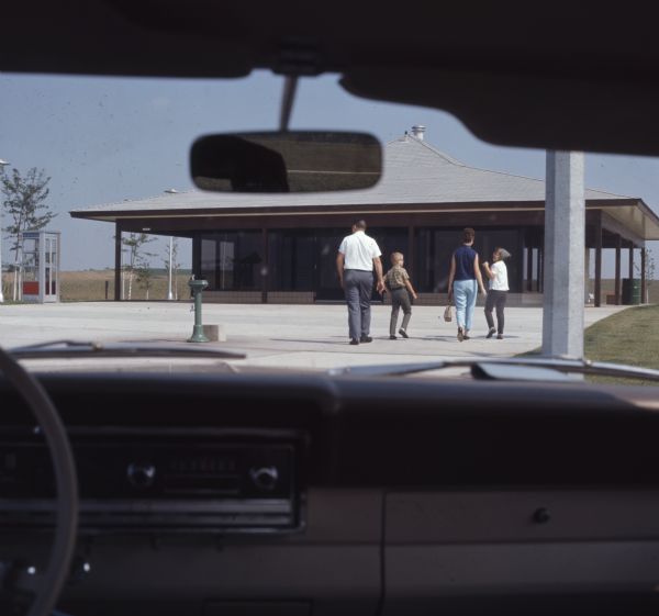 View through a car windshield towards the Beloit tourist center. A family of four is walking towards the building. A phone booth is nearby on the left.