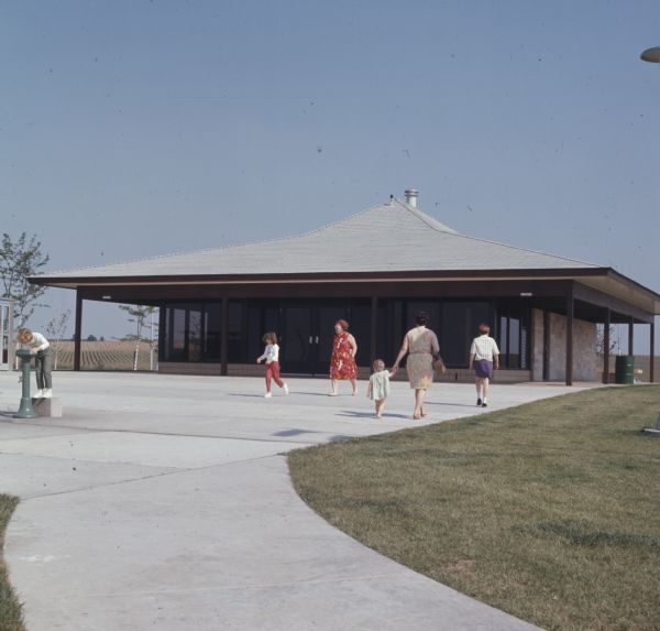 View of the Beloit Tourism Center. Women and children are walking to and from the building. A child is drinking from an outdoor water-fountain (bubbler) on the far left. 