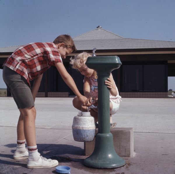 A boy and a girl are filling a jug of water from the water-fountain (bubbler) outside of the Beloit tourism center.