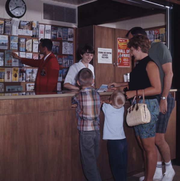 A family is standing at the desk of a tourist center. A woman behind the counter is showing them a map. Behind the counter a man is reaching for another brochure from a display.