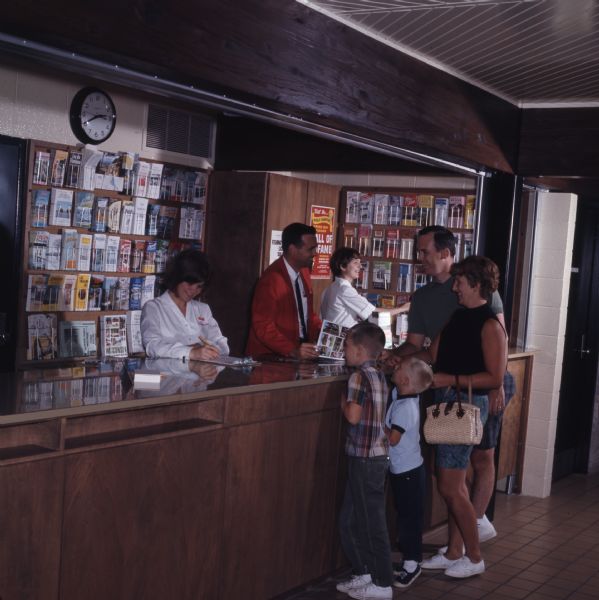Two women and a man are standing behind the counter at the Beloit Tourism Center. The man is showing a family of four a brochure. Brochures and maps are on display in racks behind the counter.