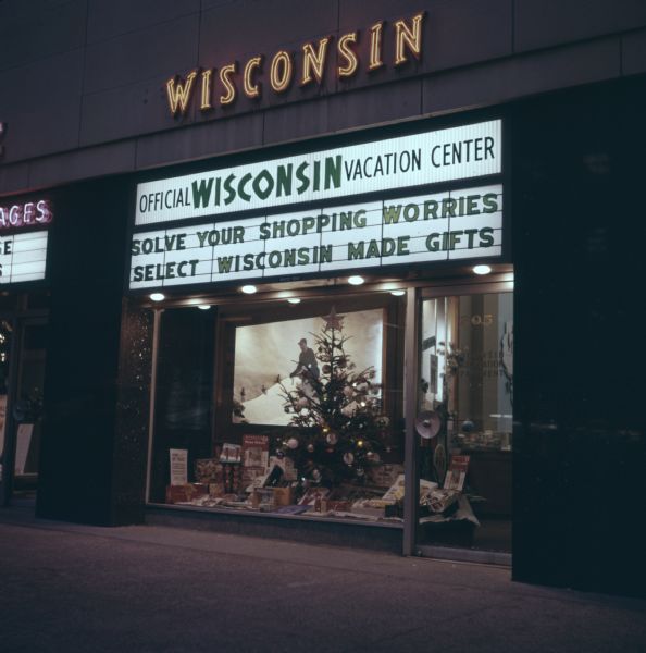 View of a storefront at night. The electric sign above the glass door and window reads: "Official Wisconsin Vacation Center: Solve Your Shopping Worries, Select Wisconsin Made Gifts." The sign on the glass door reads: "Wisconsin Conservation Department." Above the show window there is a sign that reads: "Wisconsin" above the store front in neon lights. Books, gifts, a large image of a man on a snowy landscape, and a christmas tree all are in the window display.