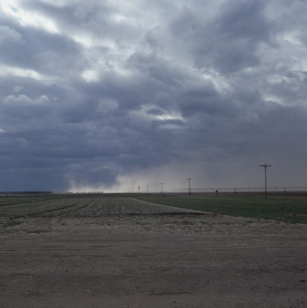 A dust storm is rising up across a field. A center pivot irrigation system is on the right in the distance beyond power poles.