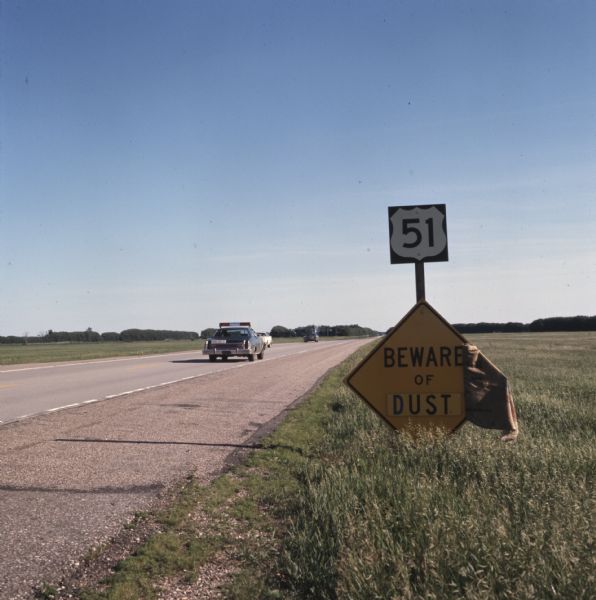 View from side of road towards the rear of a police car driving down U.S. Route 51. Signs on the side of the road read: "51" and "Beware of Dust."