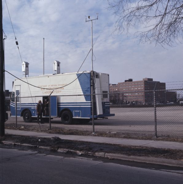 View across street through chain-link fence towards a van labeled: "Department of Natural Resources Air Monitoring Laboratory." Behind the van and across a parking lot is the Oscar Mayer & Co. headquarters building.