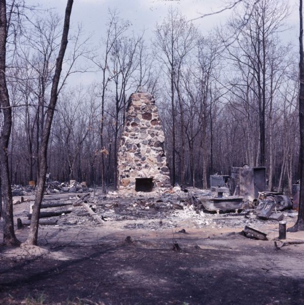 Scorched metal, ash, a foundation, and a brick chimney are all that remain of a cottage after a fire. Bare trees surround the ruins of the cottage.