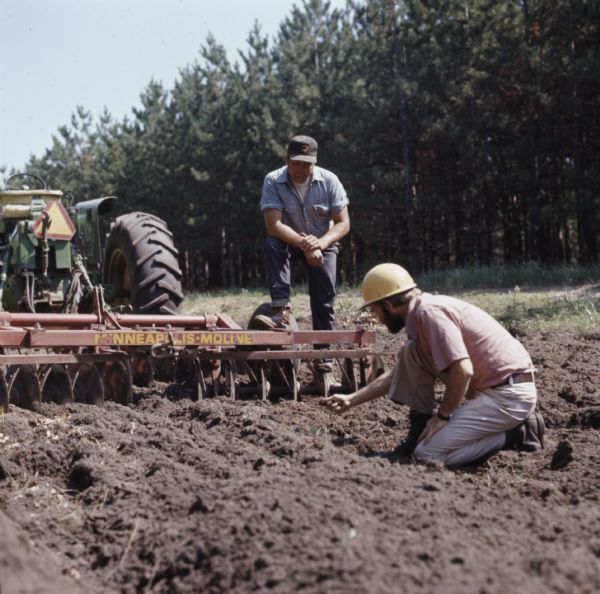 One man, wearing a hard hat, is kneeling in the dirt in the foreground, while another man wearing a cap is resting one foot on a Minneapolis-Moline disk plow attached to a tractor and looking down at the plowed area. The men are creating a fire break.