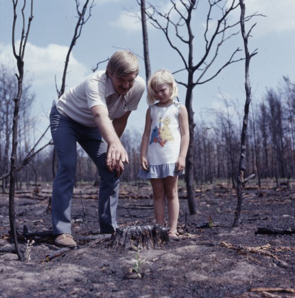 A man (Mr. Carlsen) is standing next to his daughter (Lisa). He is bending over and pointing to a small green plant growing out of the scorched ground after a forest fire.