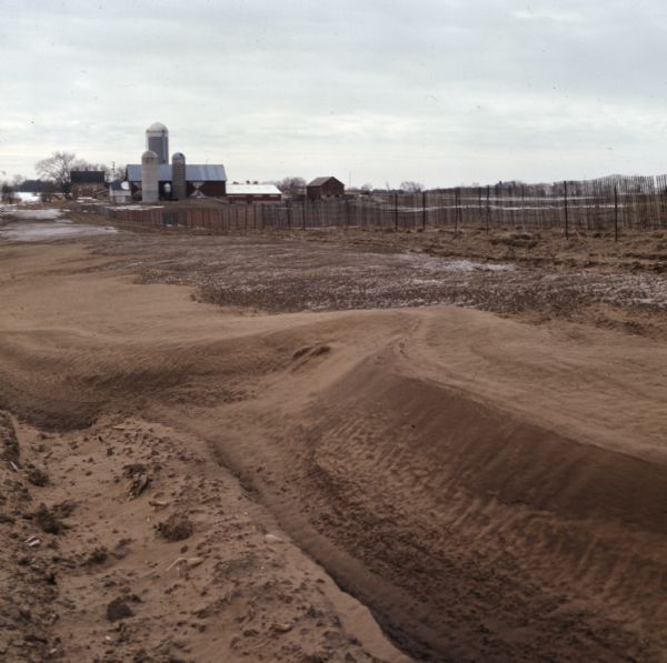 View of a dirt field showing signs of wind erosion. Farm buildings, a fence and fields are in the distance.