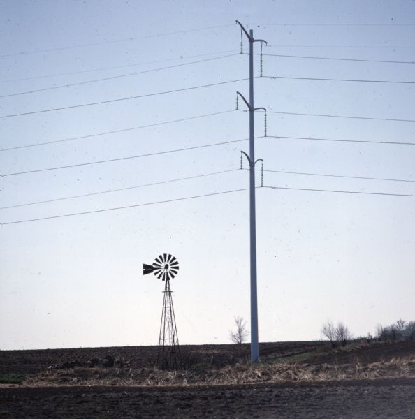View across field towards a windmill standing next to a tall electric utility pole on a field.