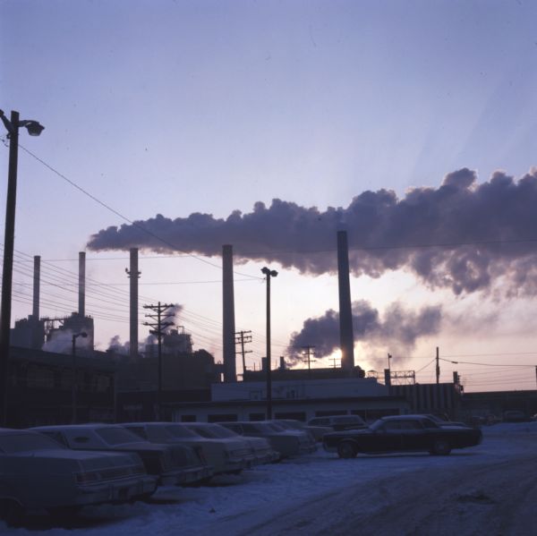 Silhouetted view of the smokestacks, buildings, and parking lot of Madison Gas & Electric at sunrise. Smoke is rising out of the smokestacks, and cars are parked in the parking lot. Snow is on the ground.