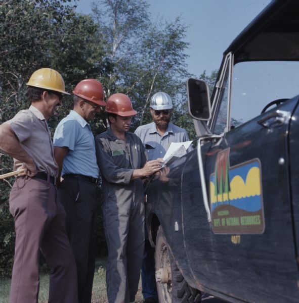 Four men of the Wisconsin's Department of Natural Resources are wearing hard hats and standing next to a truck as they look over papers. The Wisconsin DNR logo is on the driver's side door of the truck. 