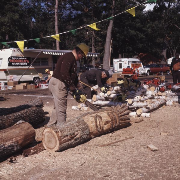 A man is carving a face onto a log using a chainsaw. Other men are looking at chainsaws displayed on a pile of birch tree logs. A sign on a trailer behind the men reads: "You're in control with Homelite Chain Saws."