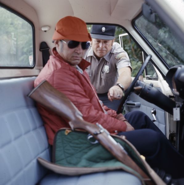 View from passenger side of truck towards a Wisconsin Department of Natural Resources warden leaning into the open door of the truck and pointing at a rifle next to the man in the driver's seat. The rifle is partially out of its case.