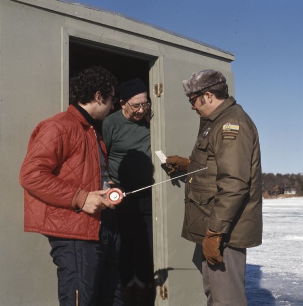 Three men are standing in or near the door to an ice fishing shack. One man is holding a fishing pole, and another man is standing in the open doorway of the ice fishing shack. The third man, wearing the uniform of a Department of Natural Resources warden, is inspecting the ice fishing license.
