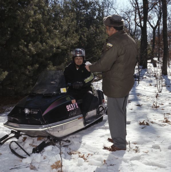 A warden of the Department of Natural Resources is checking the permit of a man sitting on a snowmobile.