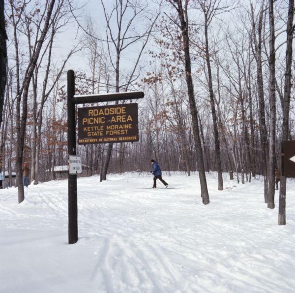 A wood sign in the foreground reads: "Roadside Picnic Area, Kettle Moraine State Forest, Department of Natural Resources." A few people are cross country skiing in the background.