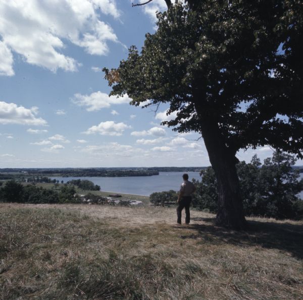 View looking down hill towards a Department of Natural Resources employee standing under a tree which is overlooking Pike Lake State Park. Pike Lake is in the background.