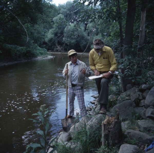 Two men are standing on the rocky shoreline of the Sheboygan River. One man is holding a shovel in his right hand and a glass jar in his left hand. The other man is writing in a notebook braced on his knee. They are measuring the PCB levels in the water.