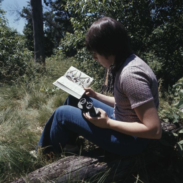 A woman is sitting on a log outdoors. She is holding binoculars in her left hand, and is reading a bird identification book.
