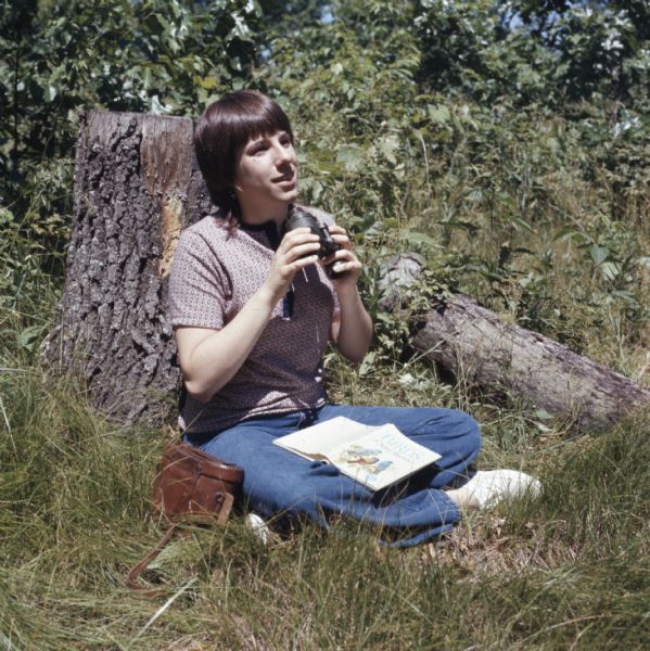 A woman is sitting in the grass next to a tree trunk. She is holding a pair of binoculars in her hands, and has a book <i>Birds of North America</i> lying in her lap.