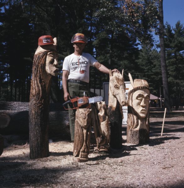 A man is posing with a number of his wood carvings. He is wearing a shirt and face-shield advertising Echo chainsaws. A chainsaw is sitting on top of a log carved into an owl. 