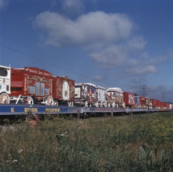 View across field towards a train of open railroad cars carrying circus wagons.