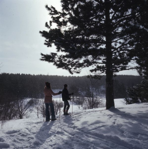 Two women are cross country skiing through the snow at the top of a hill.