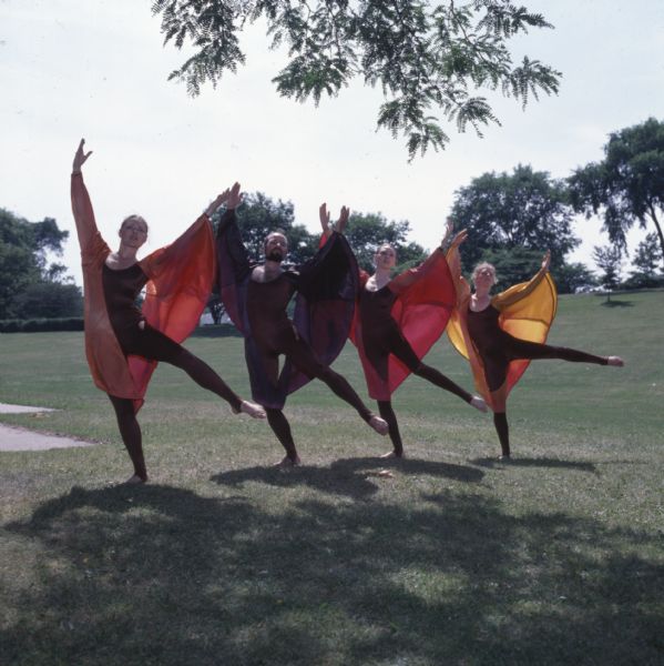 Three women and one man are standing in a line outdoors, with their arms upraised and their left legs lifted off the ground. They are wearing brown, full-body leotards and brightly colored wing-like sleeves.