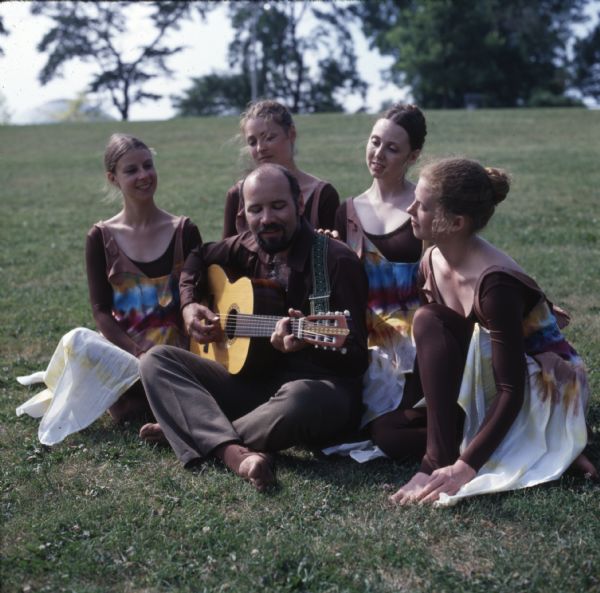 Four women are sitting outdoors in the grass around a man who is playing guitar. The women are wearing full-body brown leotards, with a tie-dye sleeveless dress over their leotards.