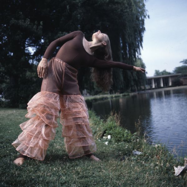 A dancer is standing on the grass near water, stretching her arm and torso to the side. She is wearing a full-body brown leotard, with ruffled, pink transparent pants. 