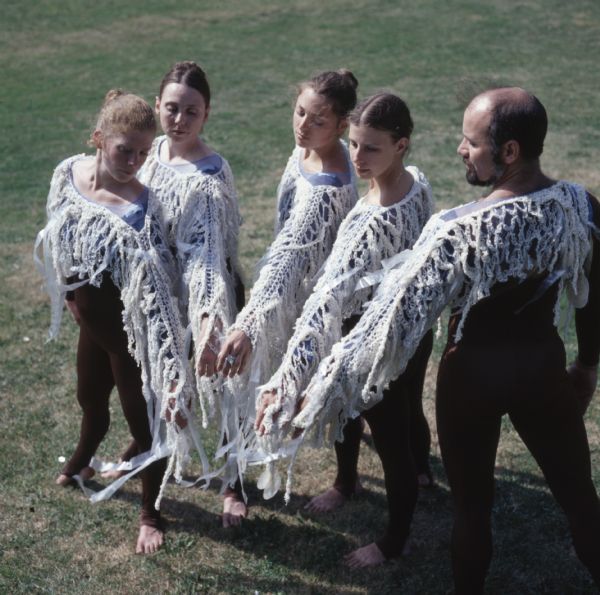 Elevated view of four women and one man standing in a semi-circle with their left arms extended inward. They are wearing full-body brown leotards, and white knit tops with the sleeves decorated with streamers.