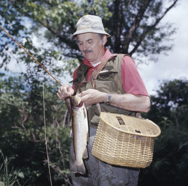 A man wearing waders, a fishing vest and a creel is holding a trout while dislodging the hook from its mouth.