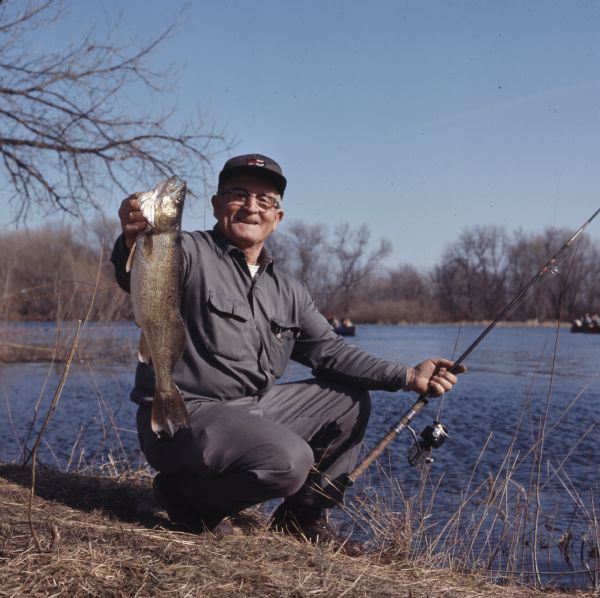 A man is kneeling on the edge of Fox River and holding his fishing pole in his left hand and a walleye in his right. Behind him other people are sitting in boats on the river.