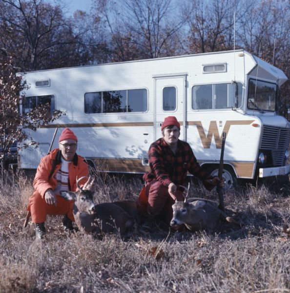 Two men wearing red and orange clothing are kneeling on the ground and posing with their bucks in front of a trailer.
