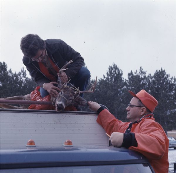At a Department of Natural Resources deer check station, a man is inspecting a buck tied to the top of a trailer. The other man is standing by the truck watching, while snow is falling around them.
